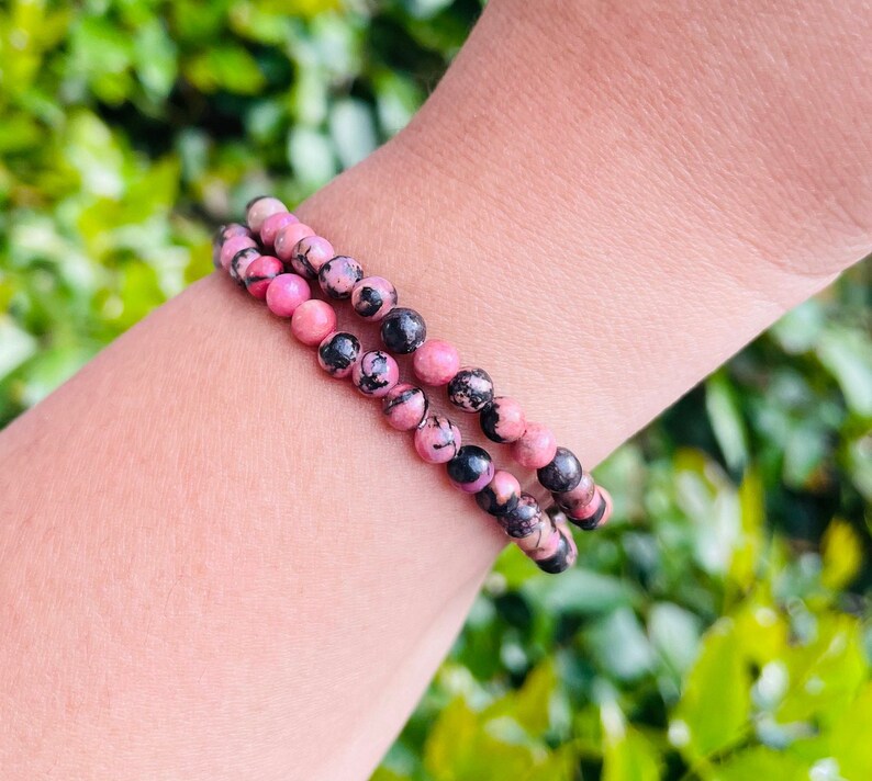 May include: Two bracelets made of pink and black gemstone beads. The bracelets are stacked on a person's wrist.
