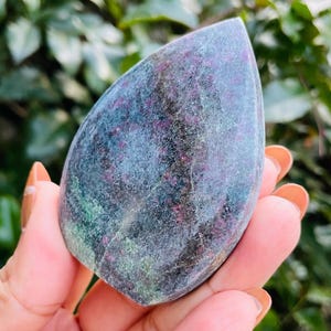 Puede incluir: Una piedra pulida en forma de lágrima con una mezcla de colores. La piedra presenta una base de azul grisáceo oscuro con parches verdes y motas rojas. La piedra se sostiene en una mano, con un fondo verde borroso.