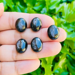 May include: Five black obsidian tumbled stones with a shiny, iridescent finish. The stones are smooth and round, and they are arranged in a row on a hand.