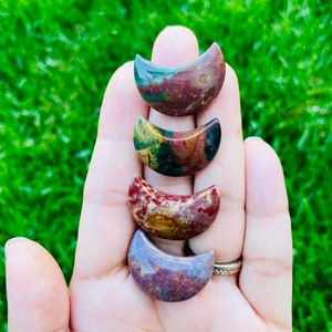 May include: Four crescent moon shaped polished stones in shades of red, brown, green, and purple. The stones are arranged in a stack, with the largest at the bottom and the smallest at the top.