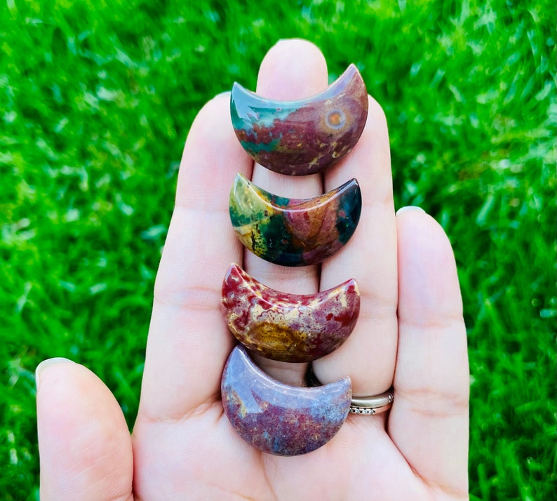 May include: Four crescent moon shaped polished stones in shades of red, brown, green, and purple. The stones are arranged in a stack, with the largest at the bottom and the smallest at the top.