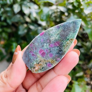 May include: A polished gemstone with a mix of colors, including deep reds, greens, and grays. The stone has a unique pattern with visible red spots and a smooth, polished surface. The gemstone is held in a hand, with a blurred green background.