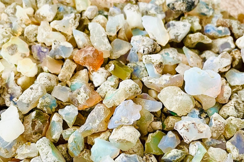 May include: A close-up of a pile of small, rough, white, and iridescent opal stones. The stones are various shapes and sizes, and some have a light blue or green hue.