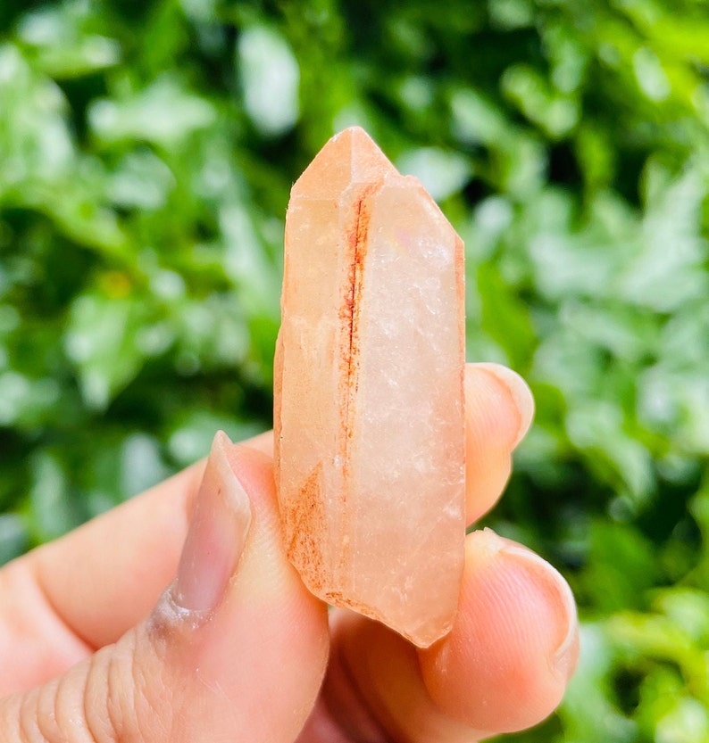 May include: A translucent, light pink crystal with a red stripe running through the center. The crystal is pointed at both ends and has a natural, rough texture.