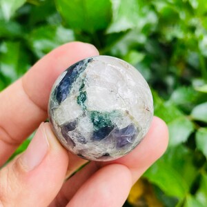 May include: A clear, spherical crystal ball with patches of dark blue, green, and white. The crystal is held in a hand, with a green leafy background.