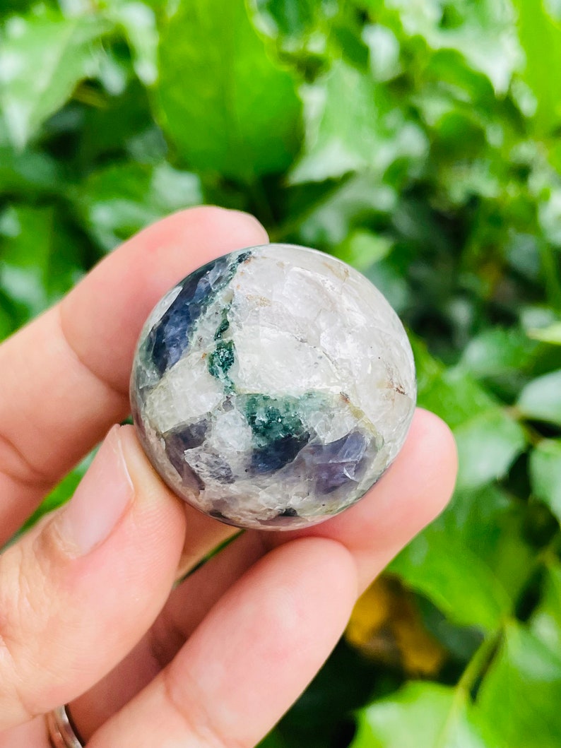 May include: A clear, spherical crystal ball with patches of dark blue, green, and white. The crystal is held in a hand, with a green leafy background.