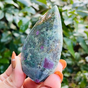 Puede incluir: Una piedra preciosa pulida en forma de lágrima, con un color base verde e inclusiones rojas vibrantes. La piedra se sostiene en una mano, con un fondo borroso de follaje verde. La superficie de la gema tiene una apariencia texturizada.