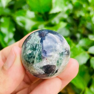 May include: A polished, spherical gemstone with a mix of green, blue, and white hues. The crystal ball is held in a hand, with a blurred green background of foliage. The sphere appears to be approximately 2 inches in diameter.