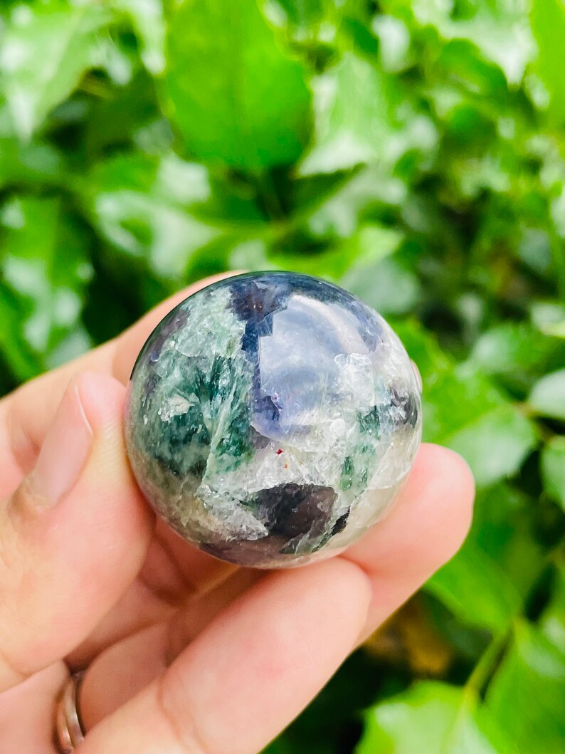 May include: A polished, spherical gemstone with a mix of green, blue, and white hues. The crystal ball is held in a hand, with a blurred green background of foliage. The sphere appears to be approximately 2 inches in diameter.