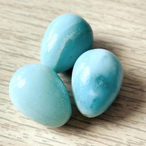 May include: Three light blue, egg-shaped stones with a smooth, polished surface. The stones have subtle white marbling and are arranged on a light brown wooden surface. The stones are a soft, pastel color.