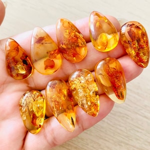 Op de afbeelding: Een verzameling van tien amber druppel hangers met insluitsels. De hangers zijn in verschillende tinten amber, van licht honing tot diep oranje. Sommige hangers hebben zichtbare insecten of plantaardig materiaal dat erin is opgesloten.