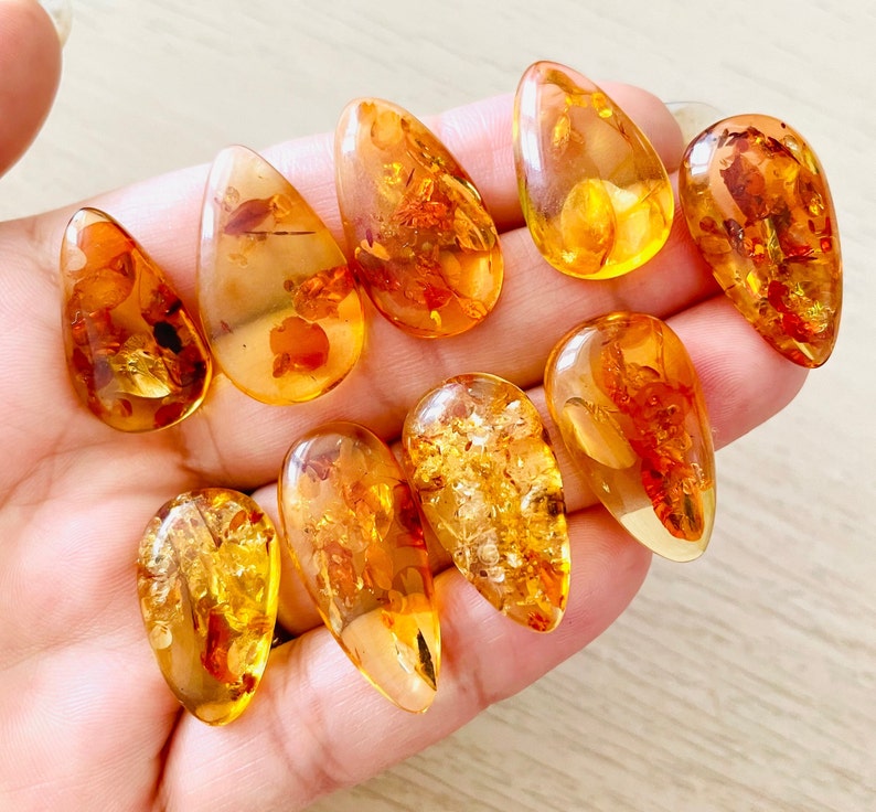 Op de afbeelding: Een verzameling van tien amber druppel hangers met insluitsels. De hangers zijn in verschillende tinten amber, van licht honing tot diep oranje. Sommige hangers hebben zichtbare insecten of plantaardig materiaal dat erin is opgesloten.
