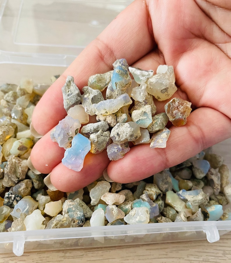 May include: A small plastic container filled with a variety of small, rough, iridescent opal stones. The stones are a mix of white, blue, and green.