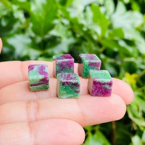 May include: Five green and pink gemstone cubes, each with a different pattern, are displayed on a hand. The cubes are approximately 1 cm in size.