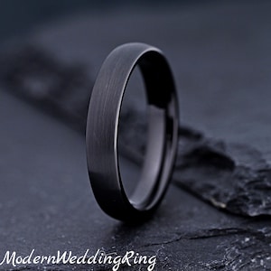 May include: A solid black ring with a brushed metal finish and a smooth, rounded profile. The band features a consistent dark tone throughout its surface, showcasing a simple, circular design that stands upright against a dark, textured surface.