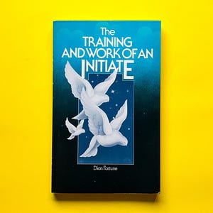 May include: A book titled "The Training and Work of an Initiate" by Dion Fortune. The cover features white doves with outstretched wings against a blue and black background. The title is in white, with the word "INITIATE" in a larger font.