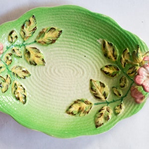 May include: A green and white oval ceramic dish with a raised design of pink roses and green leaves. The dish has a textured surface.