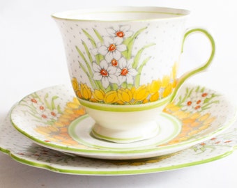 May include: A white teacup, saucer, and plate set with a floral design. The teacup and plate feature a pattern of white flowers with orange centers and yellow accents. The set has a green trim and a polka dot background.