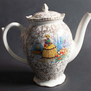 May include: White ceramic teapot with a gold floral pattern and a painted scene of a woman in a yellow dress and a blue hat. The teapot has a lid with a scalloped edge.