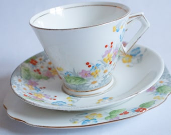 May include: A white teacup, saucer, and plate set with a floral design. The cup has a geometric handle and gold trim. The saucer and plate feature a gray border with colorful flowers and berries. The set is ideal for serving tea.