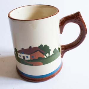 Può includere: Tazza in ceramica color crema con manico e bordo marrone. La tazza presenta un paesaggio con una casa, alberi e una fascia verde, marrone e blu attorno alla base. Il manico ha un ornamento decorativo.