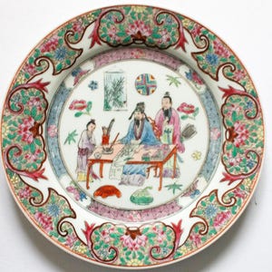 Puede incluir: Un plato decorativo de porcelana con un diseño detallado pintado a mano. El plato presenta una escena central de figuras alrededor de una mesa, rodeada por un borde floral en tonos de rosa, verde y azul. El plato tiene un fondo blanco.