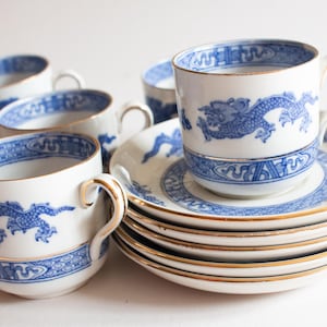 Op de afbeelding: Een set witte porseleinen kopjes en schotels met een blauw drakenontwerp. De kopjes en schotels hebben een gouden rand. De kopjes hebben oren. De set bevat meerdere kopjes en schotels.