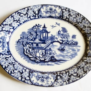 Pode incluir: Um prato oval para servir azul e branco com uma borda floral e um desenho central de uma pagoda chinesa, uma ponte e um barco.