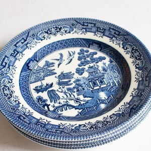 May include: A stack of blue and white ceramic plates. Each plate features a detailed Willow pattern design with a central landscape scene, including a bridge, pagoda, and birds. The plates have a decorative border.
