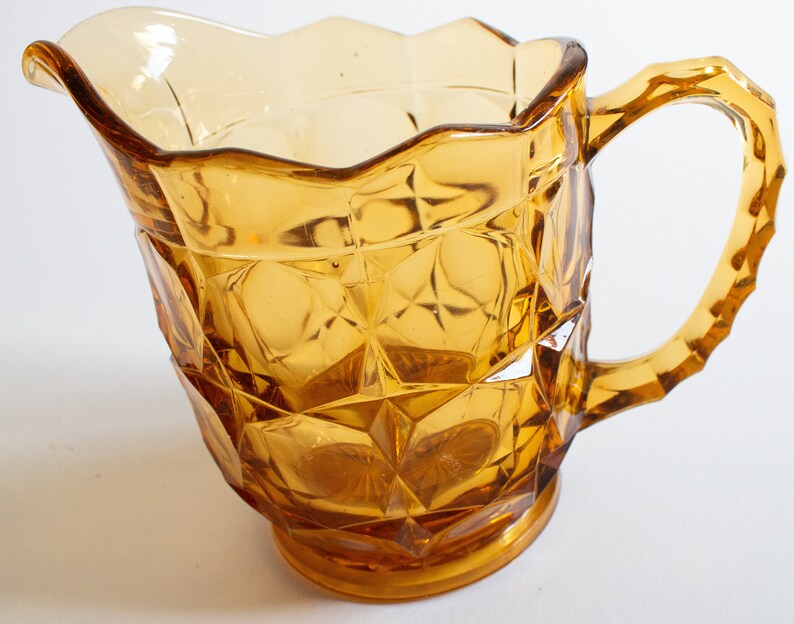 May include: Amber glass pitcher with a faceted design and a handle. The pitcher has a scalloped rim and a wide base.