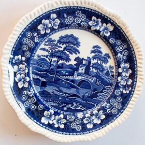 May include: Blue and white porcelain dinner plate with a scenic landscape design featuring a bridge, trees, and a stone building. The plate has a scalloped edge.