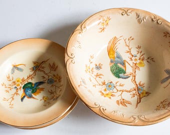 May include: A set of cream-colored ceramic bowls and plates with a bird and floral design. The bowls and plates have a gold trim and a decorative edge. The bird is blue, green, and yellow.