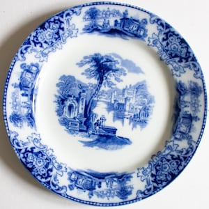 May include: A white ceramic plate with a blue landscape design. The plate features a central scene of a tree, buildings, and figures near water, surrounded by a detailed blue floral border. The plate has a classic, vintage aesthetic.