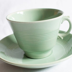 Puede incluir: Un juego de taza y platillo de té de color verde claro con un diseño sencillo. La taza tiene una forma ancha y redondeada y un asa. El platillo tiene un diseño a juego con círculos concéntricos.