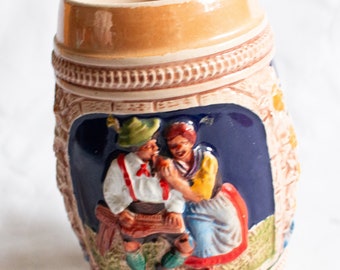 May include: A ceramic beer stein with a brown and white exterior. The stein features a raised relief design of a couple in traditional German clothing. The couple are sitting on a bench with a blue background. The stein has a brown rim and a handle.
