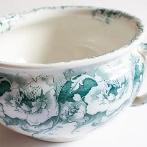 May include: A white ceramic chamber pot featuring a repeating green floral pattern around the exterior and a matching green decorative border along the rim and handle. The vessel has a wide, rounded basin and a single curved side handle.
