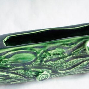 Puede incluir: Un macetero de cerámica verde con forma de tronco de árbol y superficie texturizada. El macetero tiene una abertura rectangular para plantar.