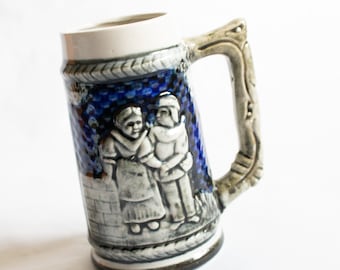 Op de afbeelding: Een blauwe en witte keramische bierkan met een reliëfdesign van twee mensen in traditionele kleding. De kan heeft een handvat en een getextureerd oppervlak.