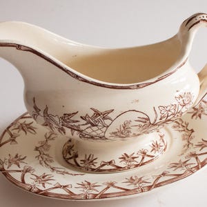 May include: Cream-colored gravy boat and saucer set with brown floral and geometric patterns. The gravy boat has a curved spout and a handle. The saucer is round with a slightly raised edge. A classic addition to any dining table.