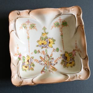 Puede incluir: Plato cuadrado de porcelana antigua con diseño floral. El plato tiene una base color crema con un borde color melocotón y ribete dorado. El diseño floral presenta flores amarillas, moradas y verdes con una cinta.