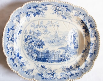 May include: A white oval serving platter with a blue and white floral design. The center of the platter features a blue and white landscape scene with a building and trees.