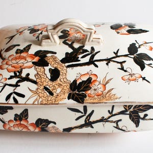 May include: Cream-colored rectangular serving dish with lid, featuring a floral and branch design in orange, black, and gold. The dish has a handle on the lid and side handles. A decorative piece for serving.
