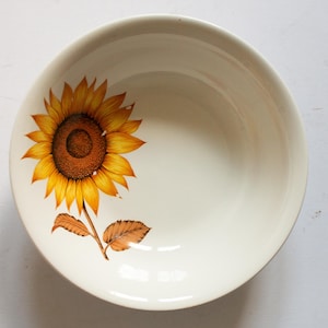 Può includere: Una ciotola in ceramica bianca con un disegno di girasole. Il girasole è giallo con un centro marrone e foglie verdi.