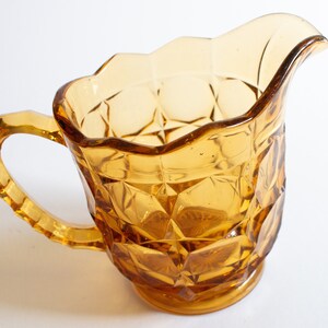 May include: Amber glass creamer with a diamond pattern and a scalloped rim. The creamer has a handle and a rounded base.