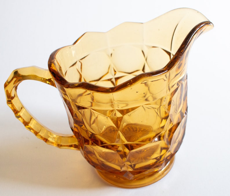 May include: Amber glass creamer with a diamond pattern and a scalloped rim. The creamer has a handle and a rounded base.