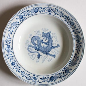 May include: White ceramic plate with a blue dragon design in the center. The plate has a blue floral border and is likely vintage or antique. The dragon is surrounded by cloud-like shapes.