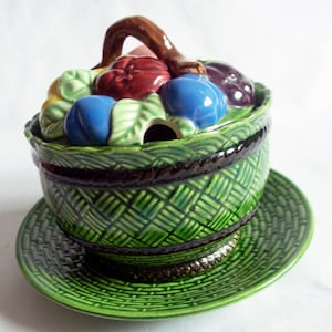 May include: A green ceramic sugar bowl with a lid shaped like a basket overflowing with fruit. The lid features a brown handle and colourful fruit, including red, blue, and purple. The bowl has a woven pattern and a matching green saucer.