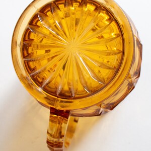 May include: Amber glass pitcher with a starburst pattern on the bottom and a handle. The pitcher has a faceted design.