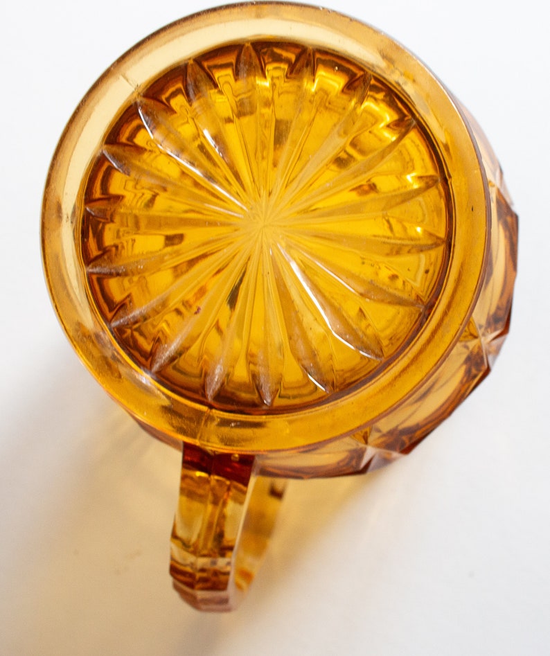 May include: Amber glass pitcher with a starburst pattern on the bottom and a handle. The pitcher has a faceted design.