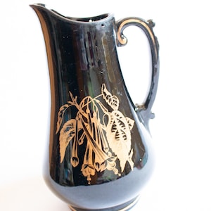 May include: Black ceramic pitcher with a gold floral design. The pitcher has a wide mouth and a handle.